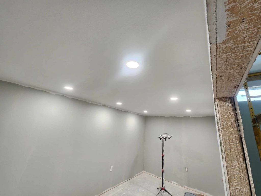 Unfinished room interior under construction with exposed drywall, recessed lighting, and repair tools visible.