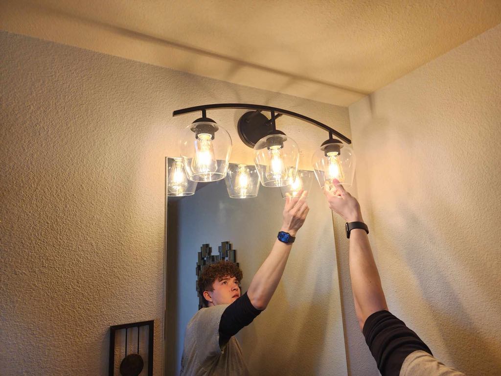 A person replacing a lightbulb in a modern bathroom vanity light fixture with five lamps.