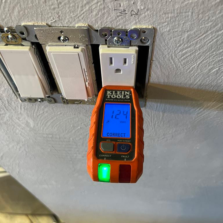 An electrical outlet tester displays 124 volts and indicates the outlet is correctly wired. The tester is plugged into a wall outlet missing its cover plate, with other switches nearby.