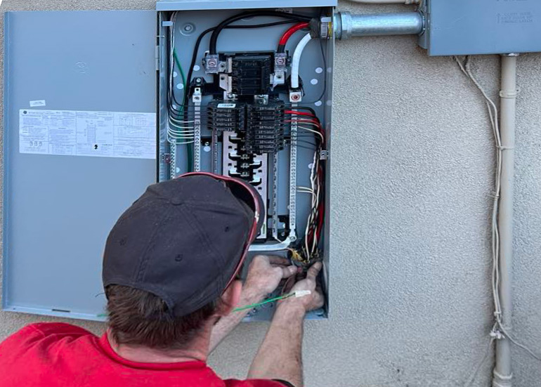 Person in a red shirt works on an open electrical panel mounted on an exterior wall, adjusting wires inside the panel while facing away from the camera.