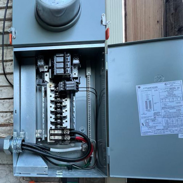 An open electrical panel with visible wiring and circuit breakers, mounted on an exterior wall. The panel door is open, revealing a label with electrical information.