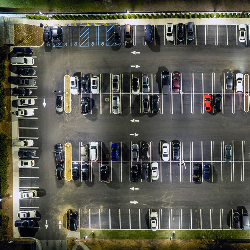 A well-lit aerial view of a parking lot at night, showing multiple rows of parked cars with clear, white parking lines and arrows directing traffic flow.