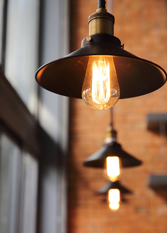 Close-up of hanging pendant lights with exposed filament bulbs against a backdrop of brick walls and large windows.