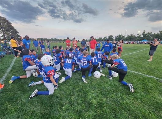 A youth football team in blue uniforms kneels on the field, listening to a coach's instructions, with spectators and trees visible in the background under a cloudy sky.